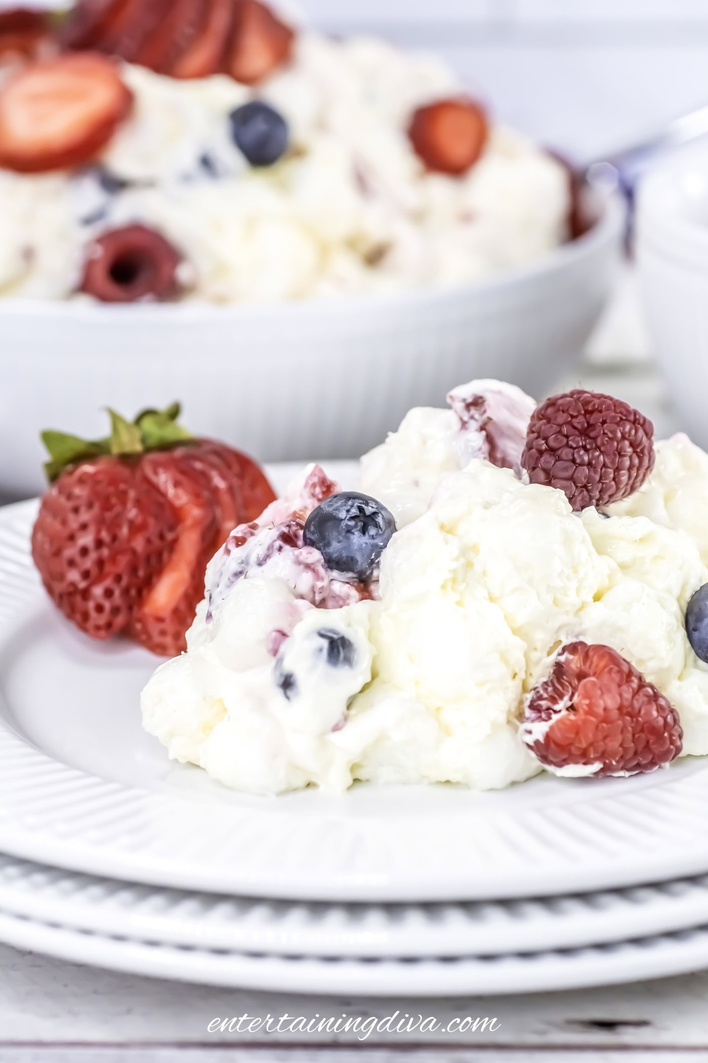 A serving of berry cheesecake fruit salad topped with raspberries, blueberries, and strawberries on a white plate, with more fruit salad in the background.