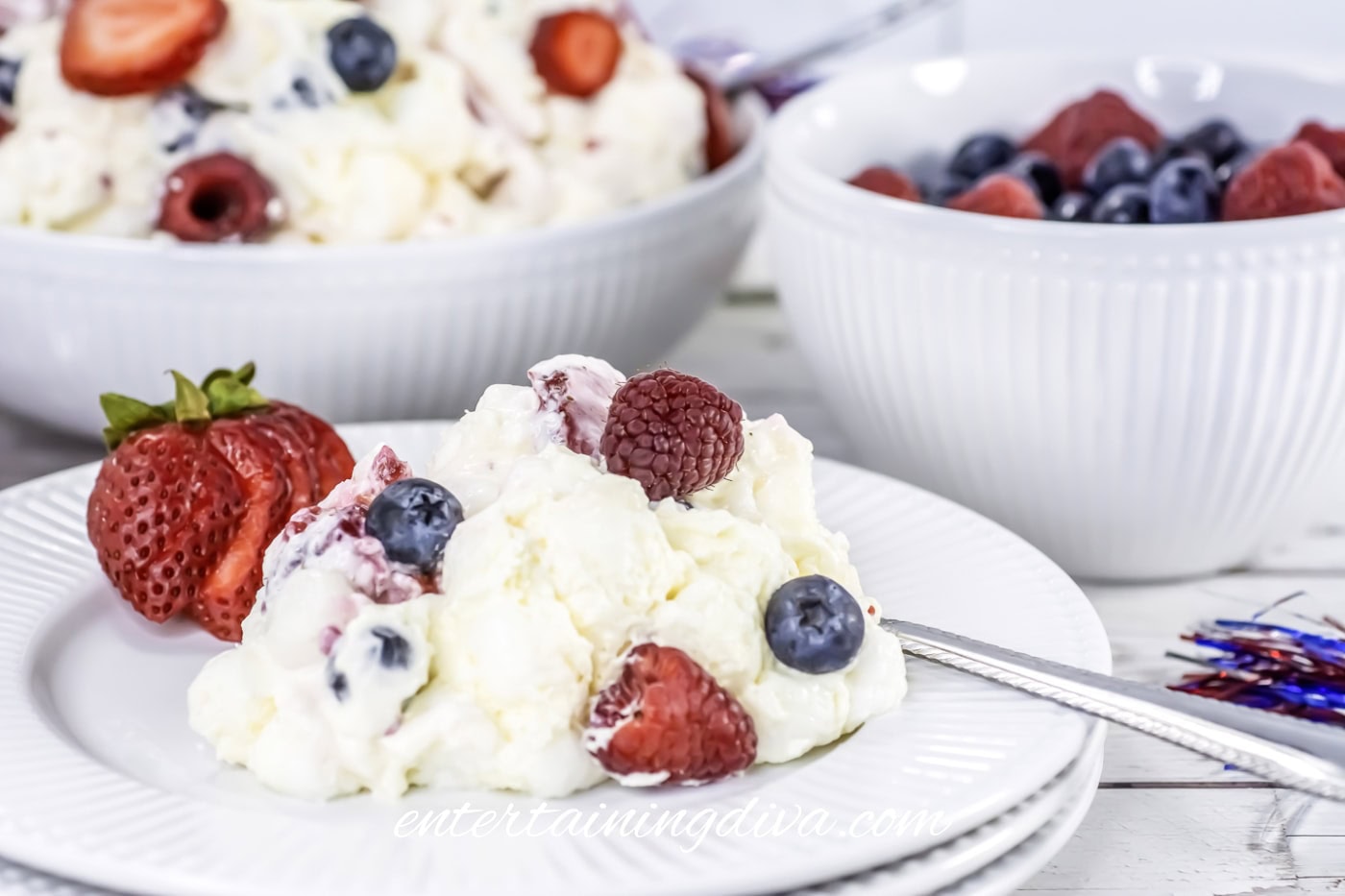 A plate of cheesecake fruit salad with strawberries, raspberries, and blueberries is served next to a bowl of mixed berries on a white table.