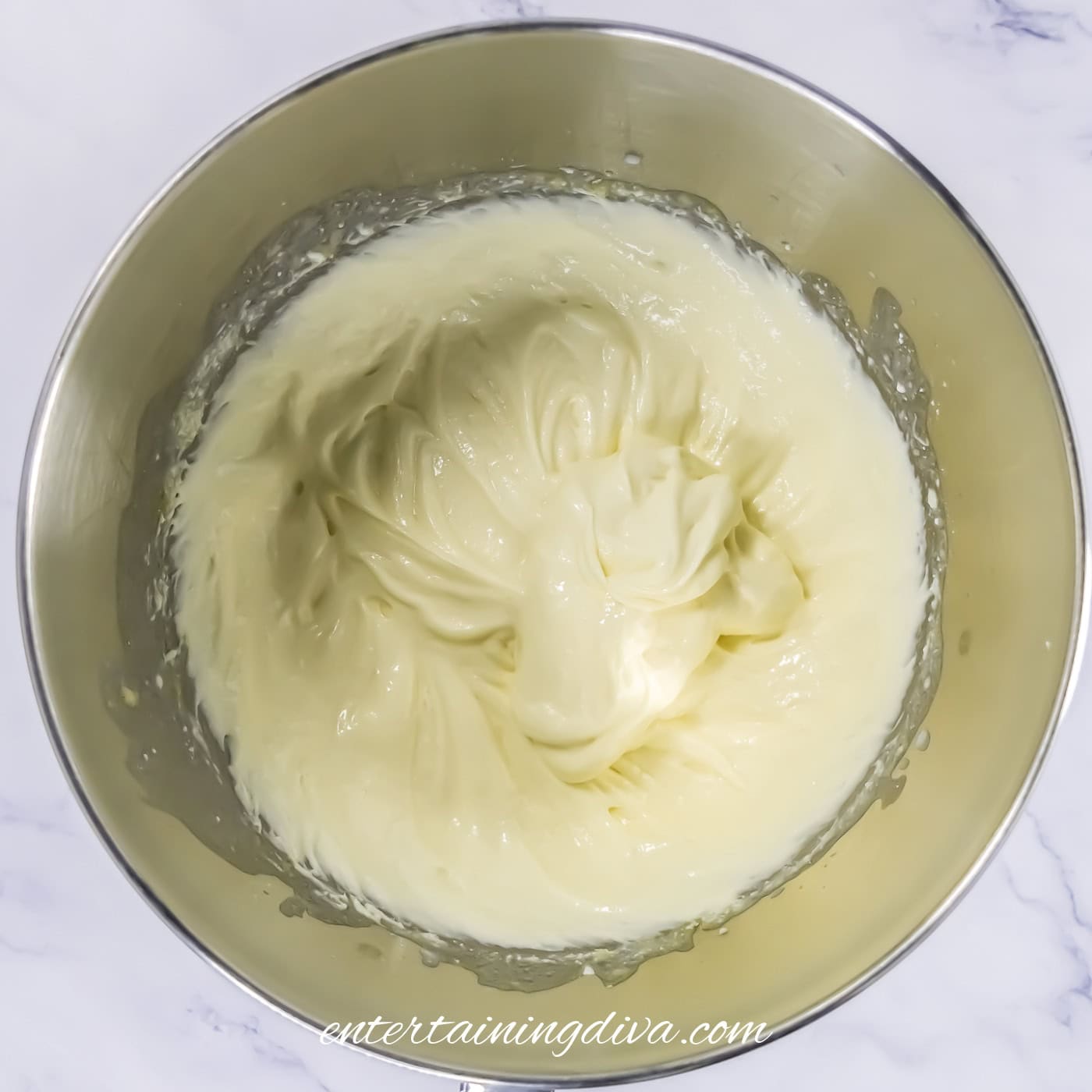 A stainless steel mixing bowl filled with a cream cheese mixture viewed from above on a white surface.