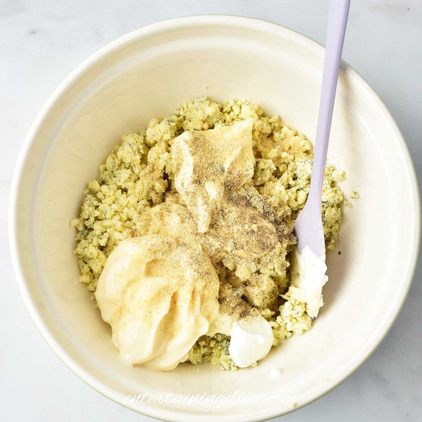 A mixing bowl containing crumbled blue cheese, Greek yogurt, mayonnaise, minced garlic and seasoning, with a spatula resting inside.
