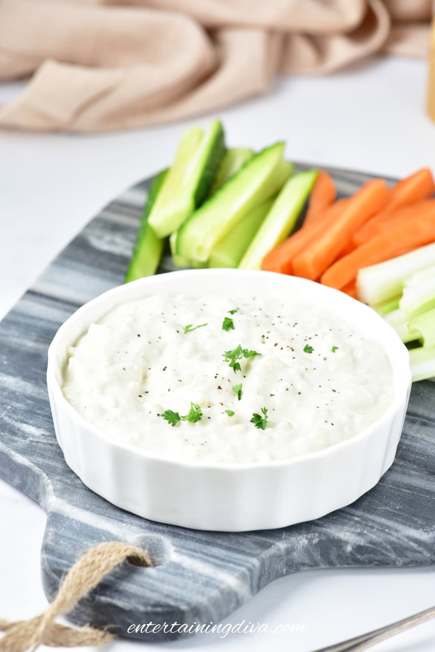 A white dish of Greek yogurt blue cheese dip garnished with herbs, served on a marble board with sliced cucumbers, carrots, and celery.