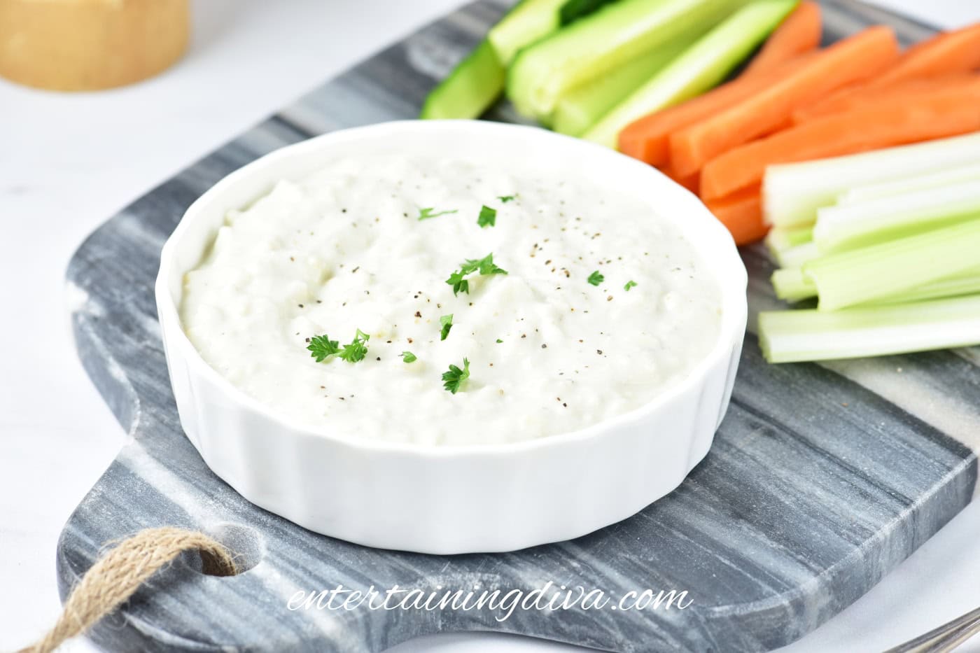 A bowl of creamy blue cheese dip garnished with herbs, served on a marble board with sliced cucumber, carrot, and celery sticks.