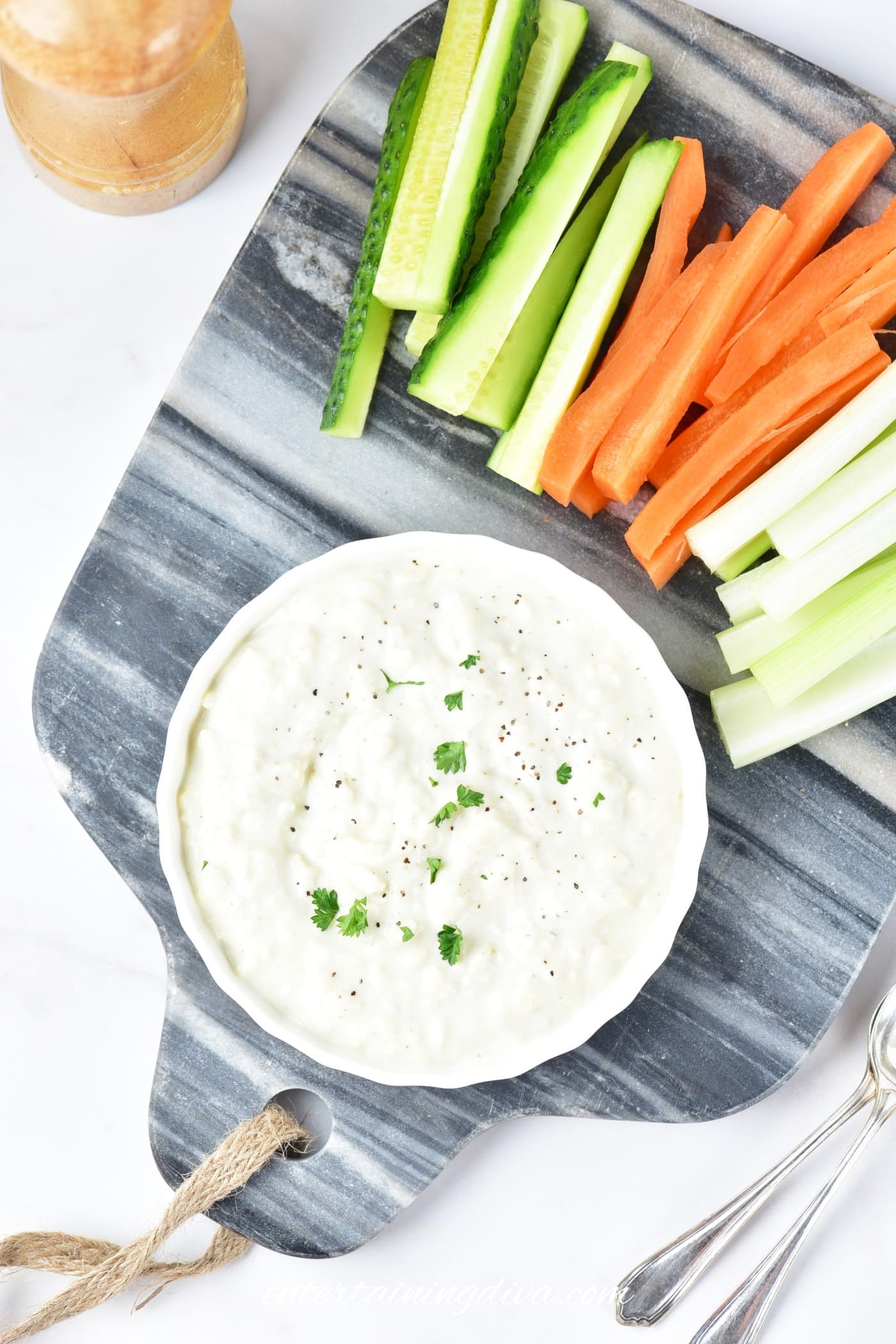 A bowl of creamy blue cheese dip garnished with herbs on a marble board next to sliced cucumber, carrot, and celery sticks.