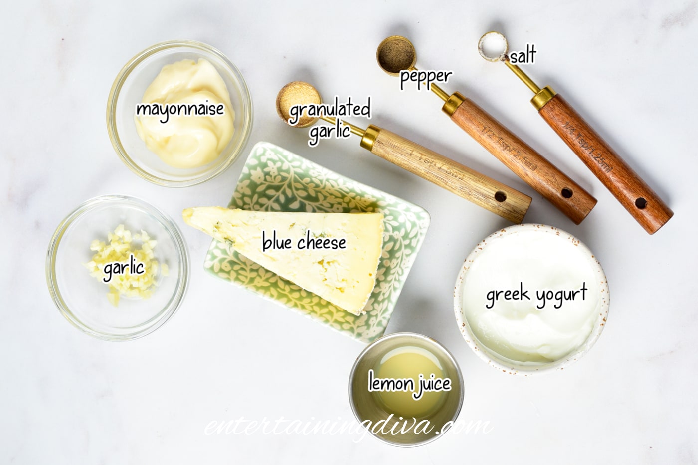 A flat lay of ingredients for a blue cheese recipe, including mayonnaise, granulated garlic, pepper, salt, garlic, blue cheese, lemon juice, and Greek yogurt in separate bowls and spoons.