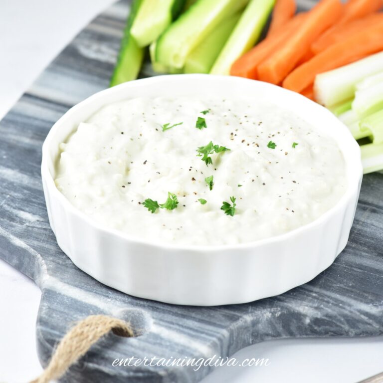 A white bowl of blue cheese dip with Greek yogurt garnished with herbs and black pepper, served on a marble board with sliced cucumbers, carrots, and celery.