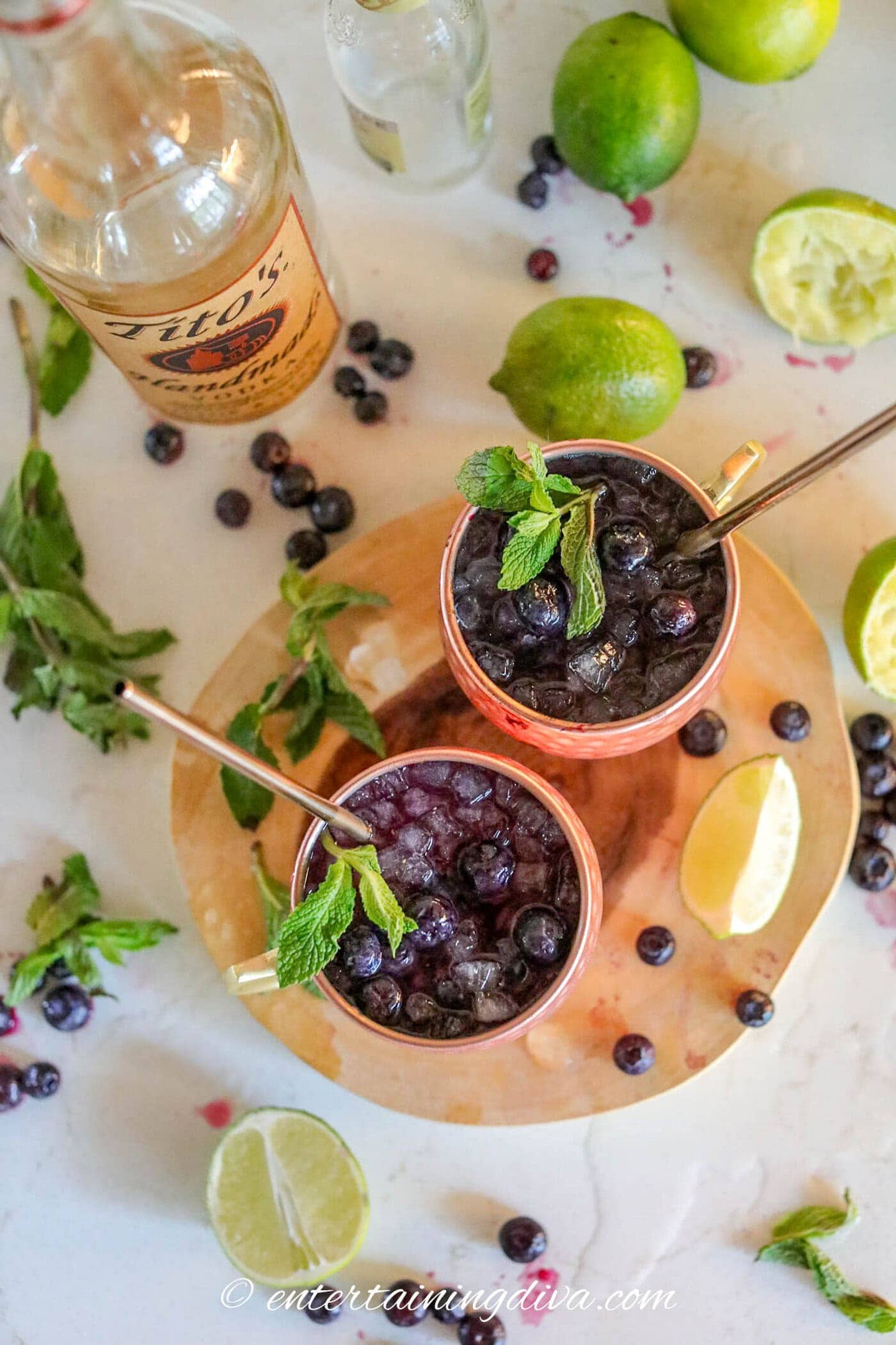 Two blueberry moscow mules in copper mugs topped with mint garnish beside a bottle of vodka