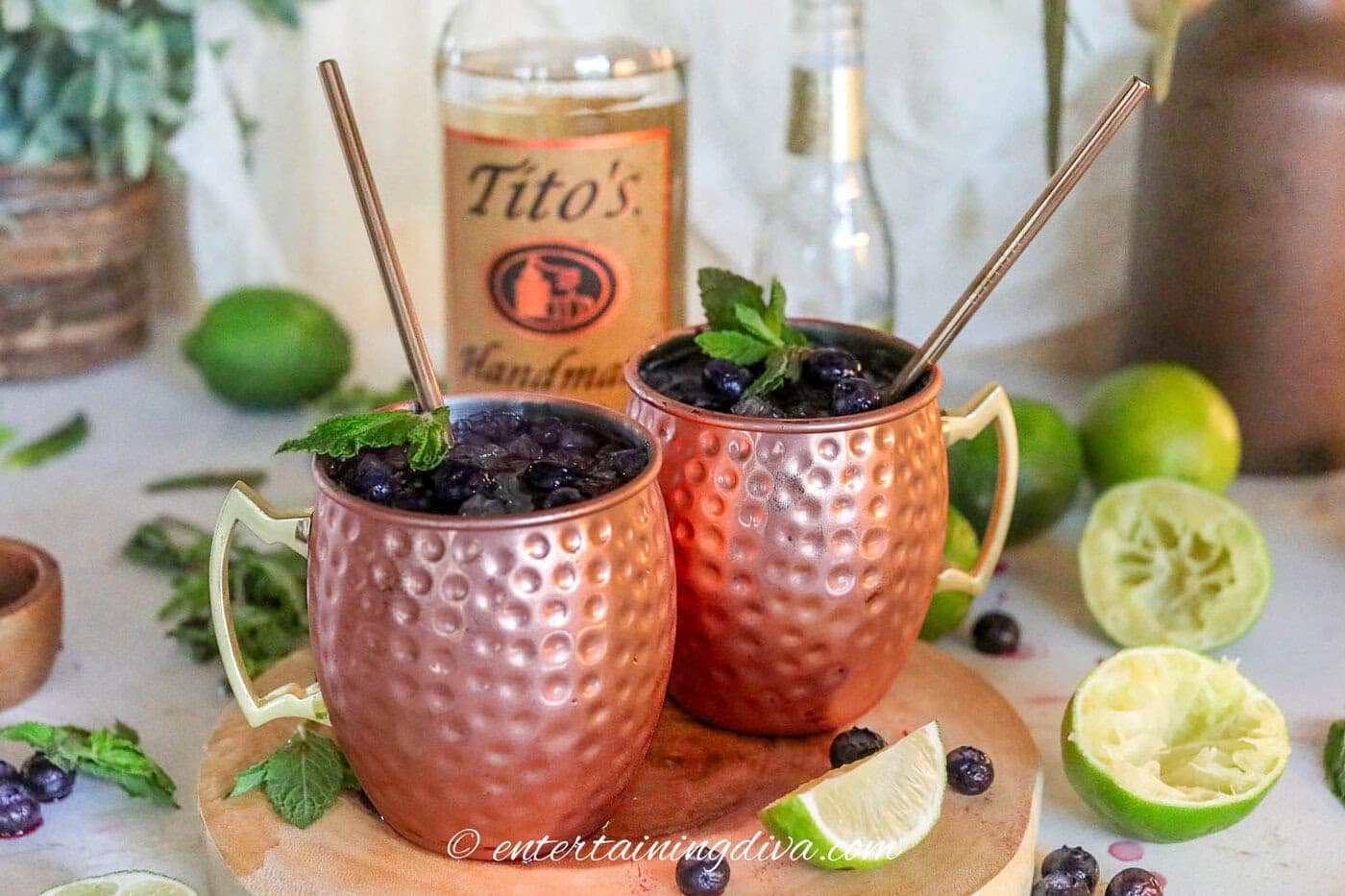 Two blueberry moscow mules garnished with mint in copper mugs in front of a bottle of vodka