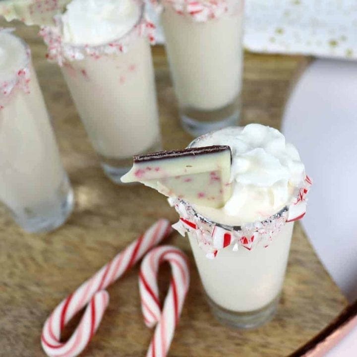 Four white festive drinks in shot glasses with whipped cream, peppermint bark, and crushed candy canes on the rims. Two candy canes lie on the wooden surface nearby.