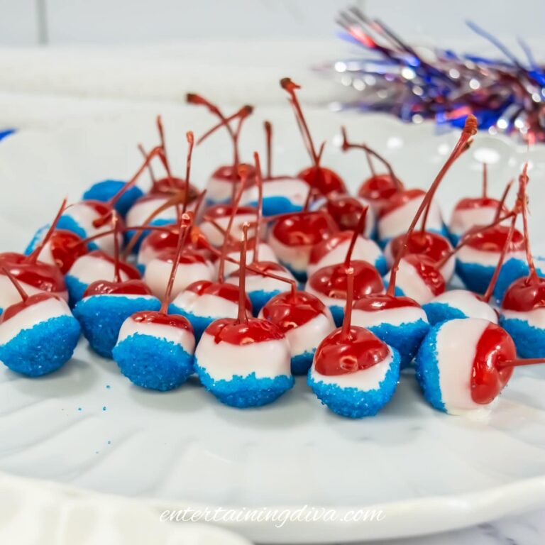A plate of cherries dipped in white chocolate and blue sprinkles, arranged neatly, with festive red, white, and blue decorations in the background.