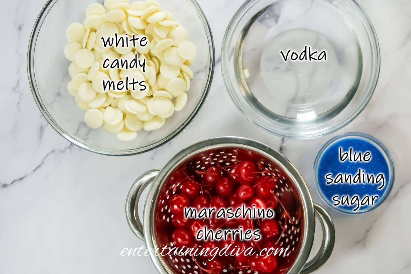 Four bowls on a marble surface containing white candy melts, vodka, blue sanding sugar, and maraschino cherries.