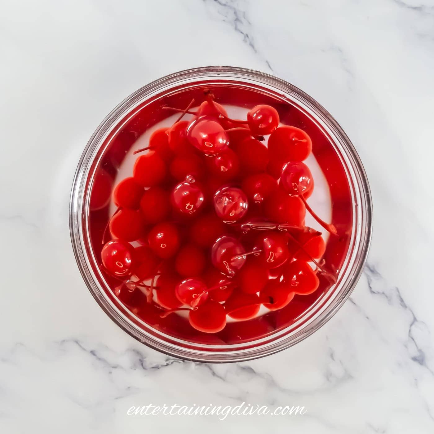 A glass bowl filled with bright red maraschino cherries in syrup, placed on a white marble surface.
