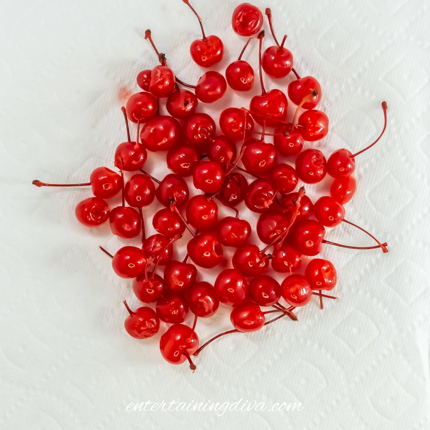 A pile of bright red maraschino cherries with stems rests on a white paper towel on a marble surface.