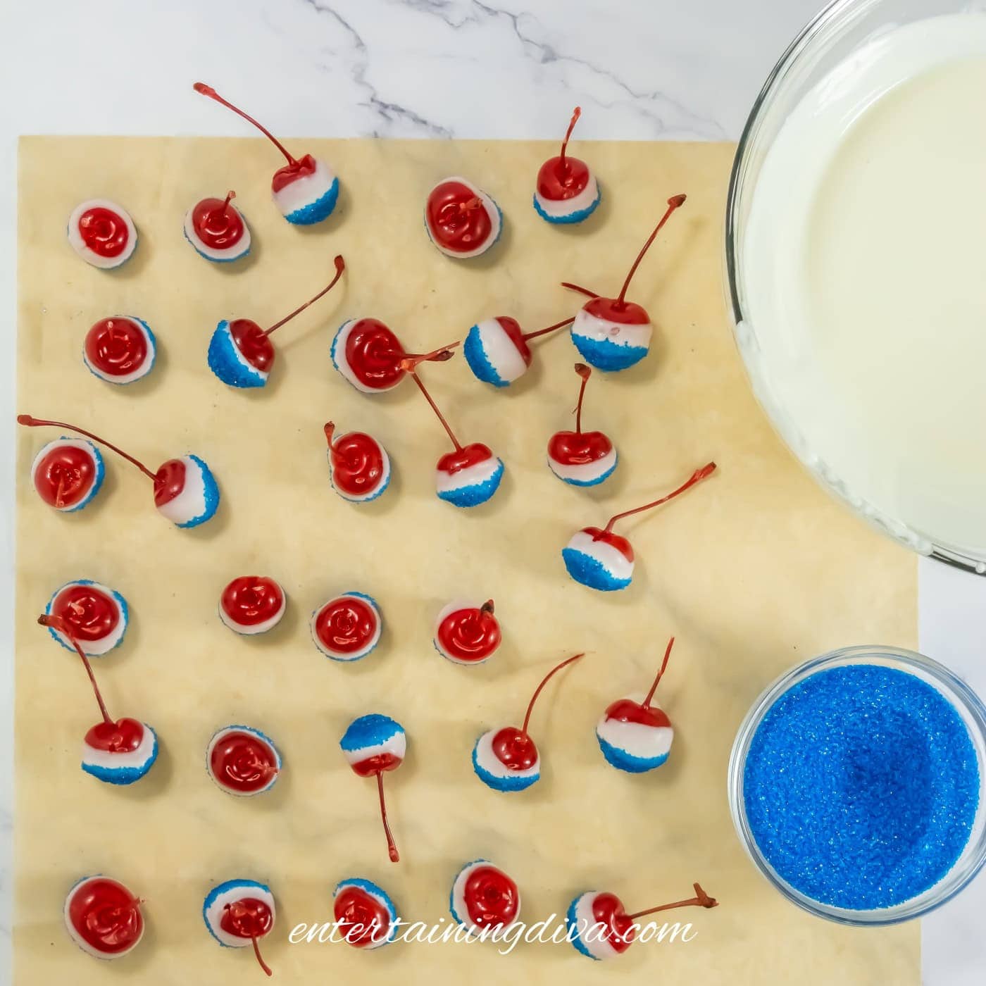 Cherries dipped in white coating and blue sprinkles arranged on parchment paper, with bowls of white coating and blue sprinkles nearby on a marble surface.