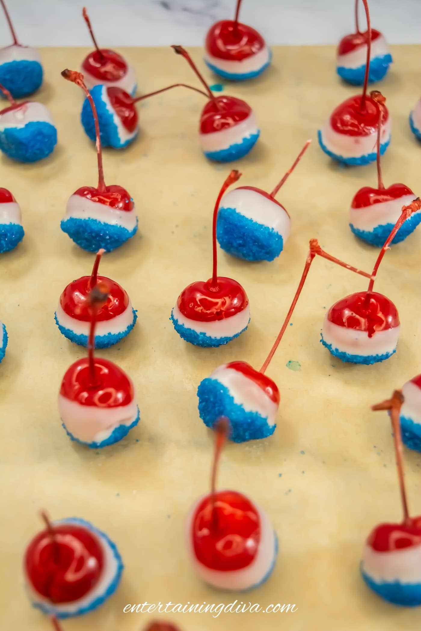 Red cherries dipped in white chocolate and blue sprinkles are arranged on parchment paper in neat rows.