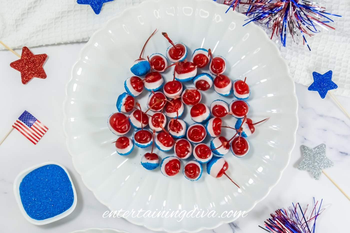 A plate of cherries dipped in blue and white coating, surrounded by patriotic-themed decorations, blue sprinkles, and mini American flags on a white surface.