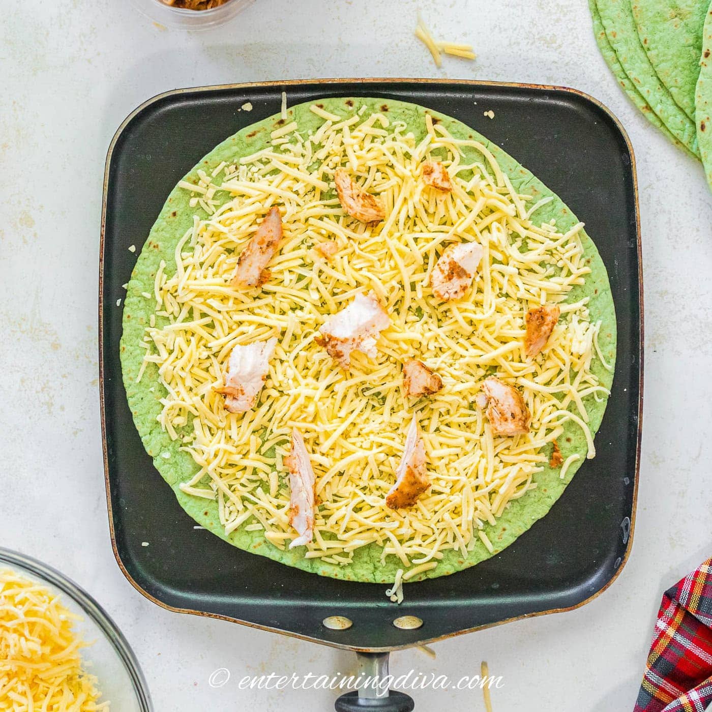 a spinach tortilla topped with grated cheese and chicken on a skillet