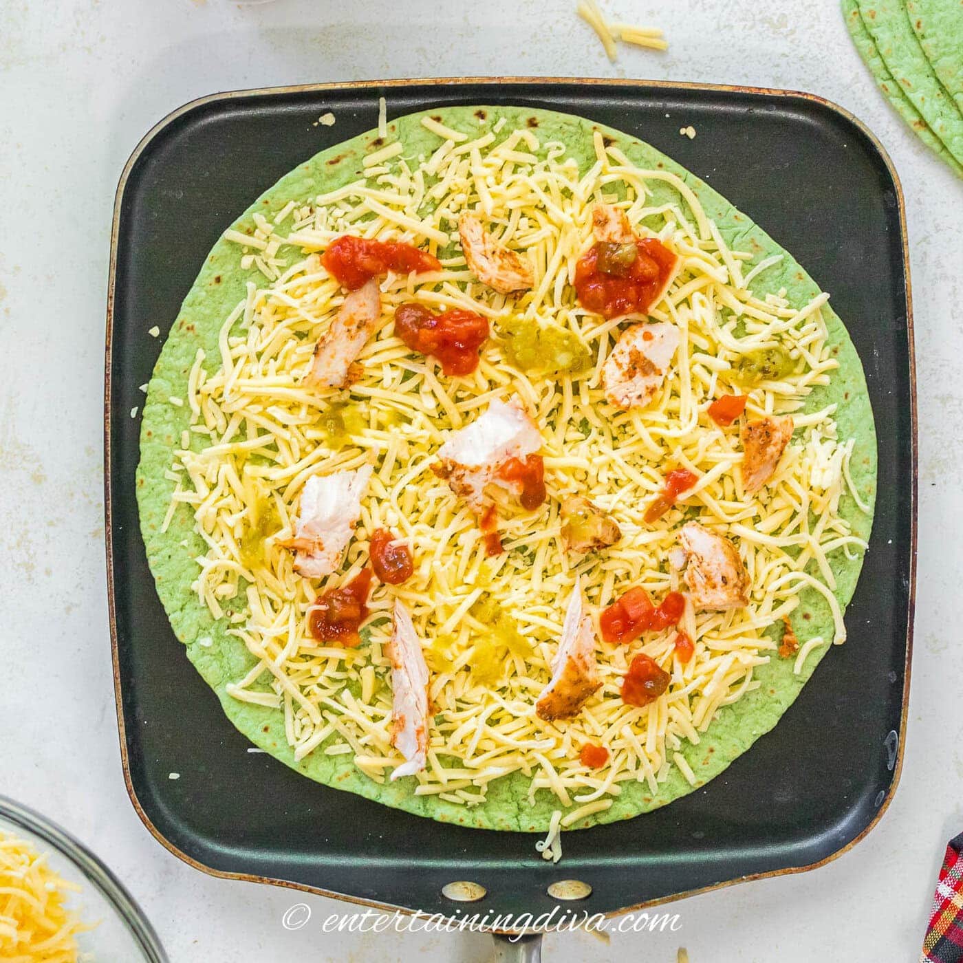 a spinach tortilla topped with grated cheese, chicken red salsa, green salsa and queso on a skillet