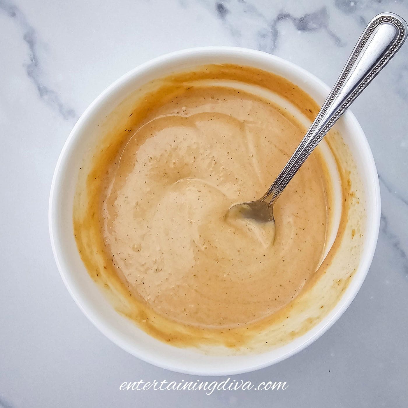A white bowl filled with a creamy, light brown dressing with a spoon inside, placed on a marble countertop.