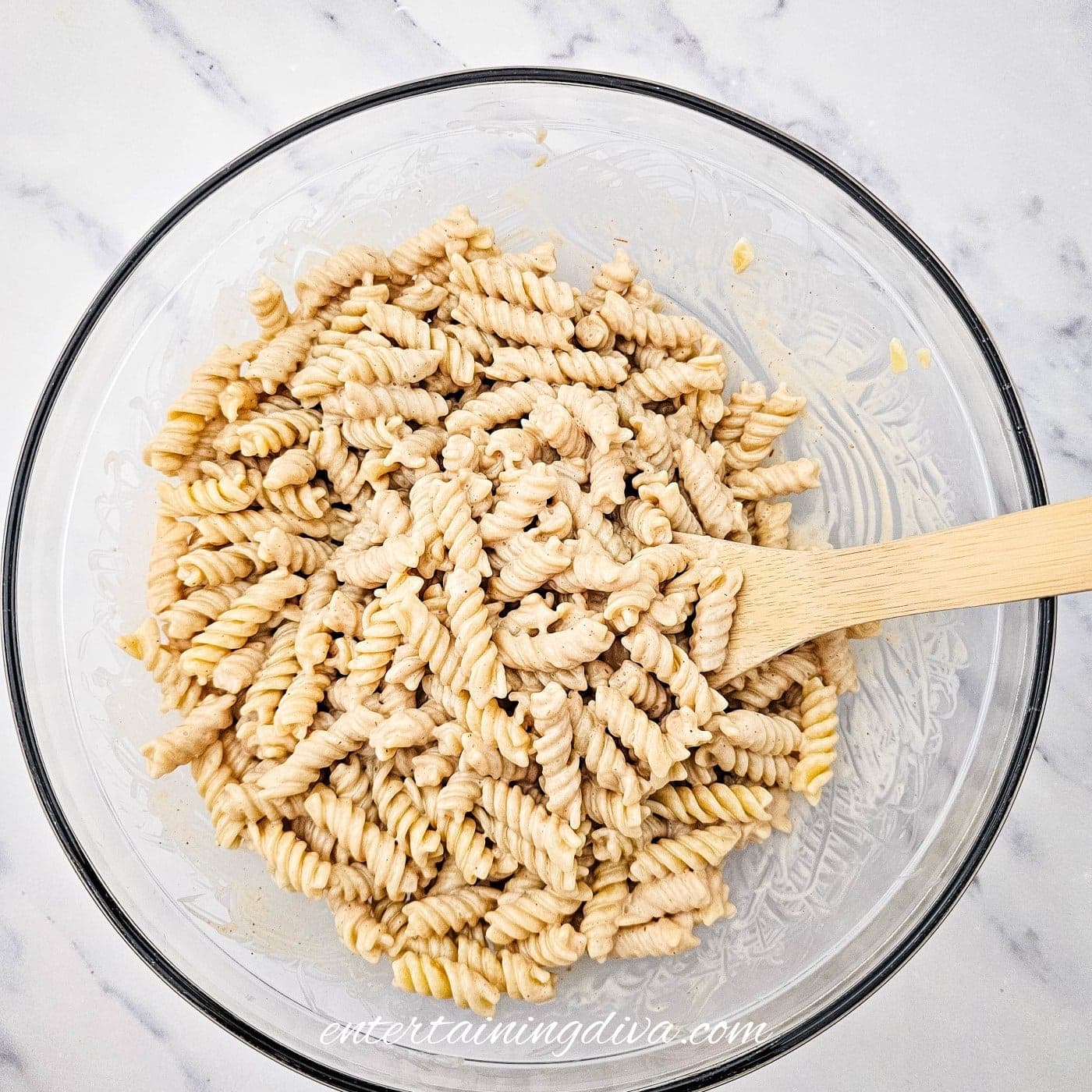 A glass bowl filled with rotini pasta mixed with a creamy sauce, with a wooden spoon resting inside the bowl on a white marble surface.