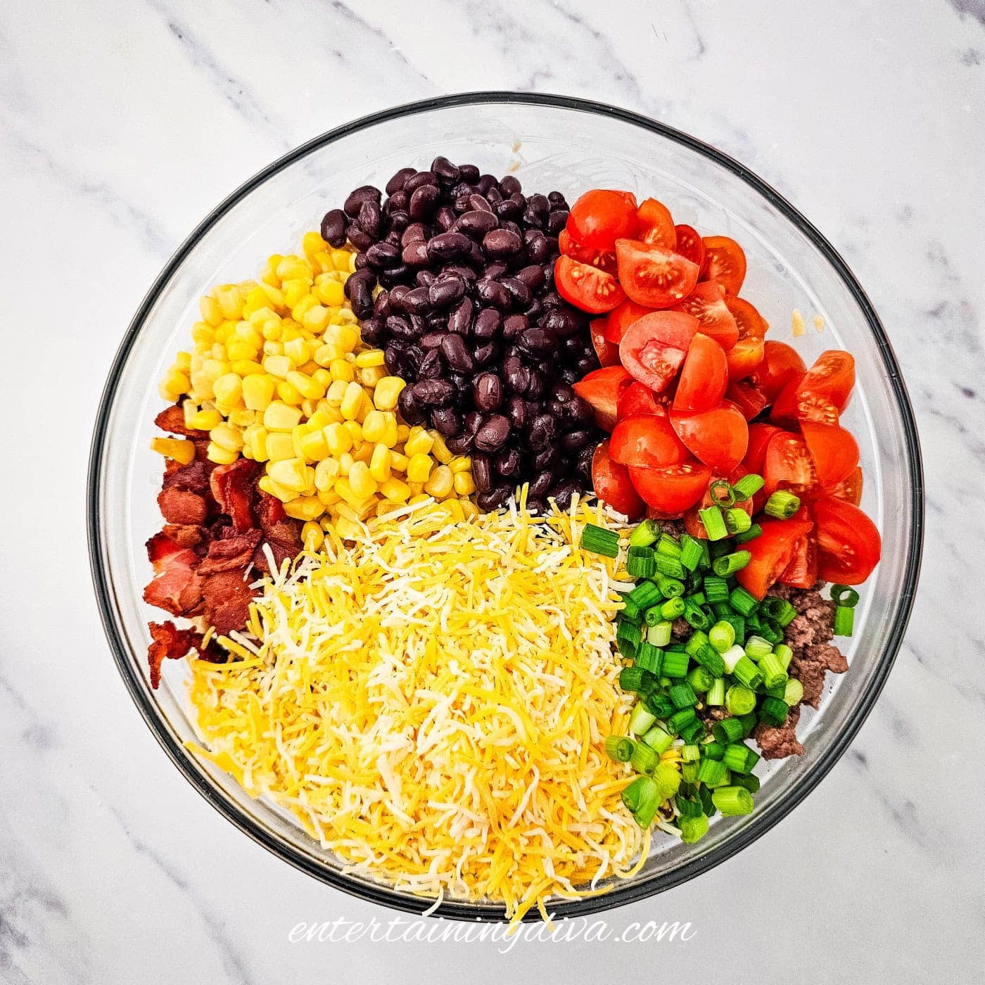 A glass bowl containing shredded cheese, chopped green onions, cherry tomatoes, black beans, corn, and bacon, arranged in separate sections on a white marble surface.