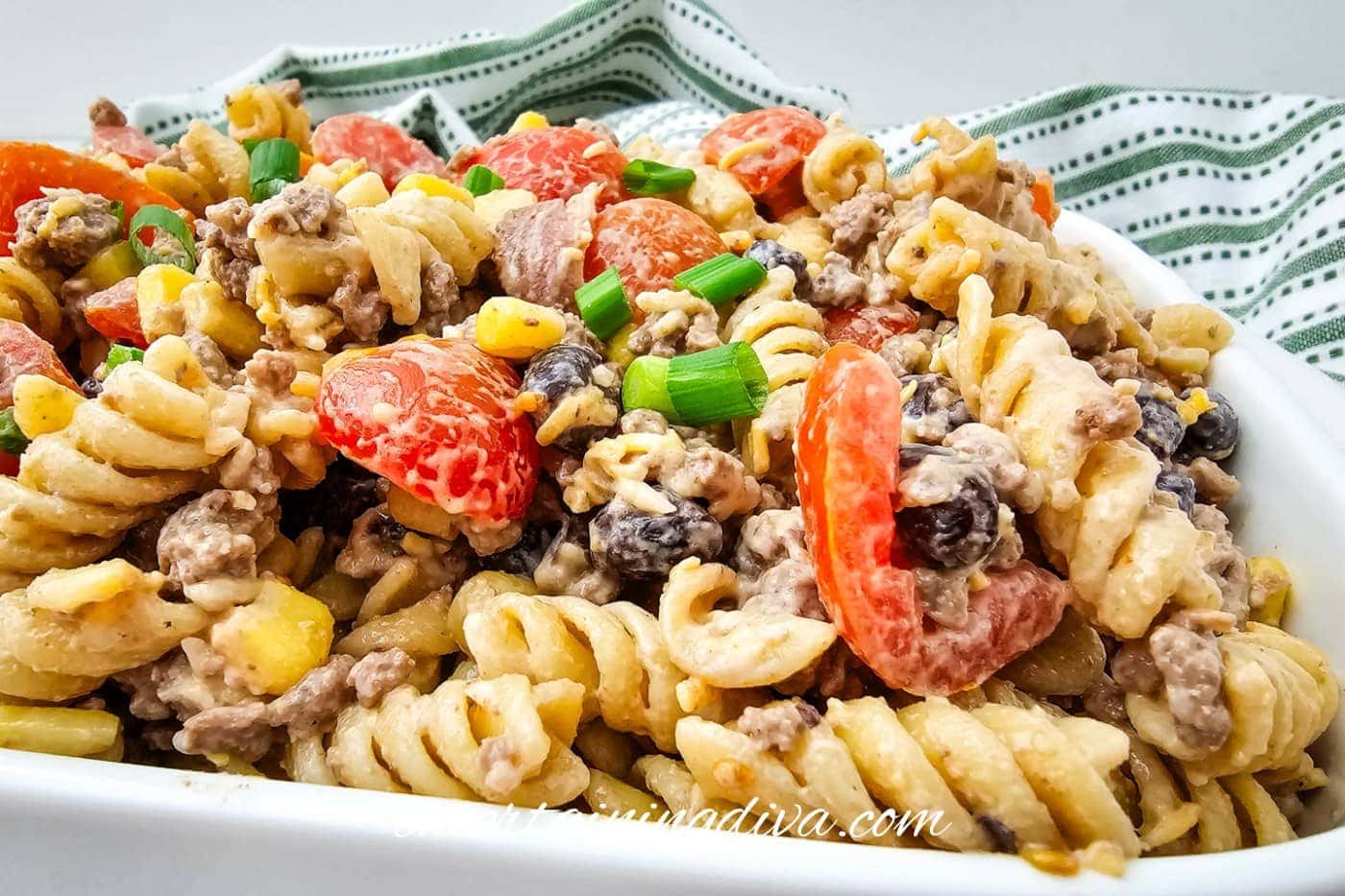 A close-up of cowboy pasta salad with rotini, ground meat, cherry tomatoes, black beans, pineapple chunks, and sliced green onions in a white dish.