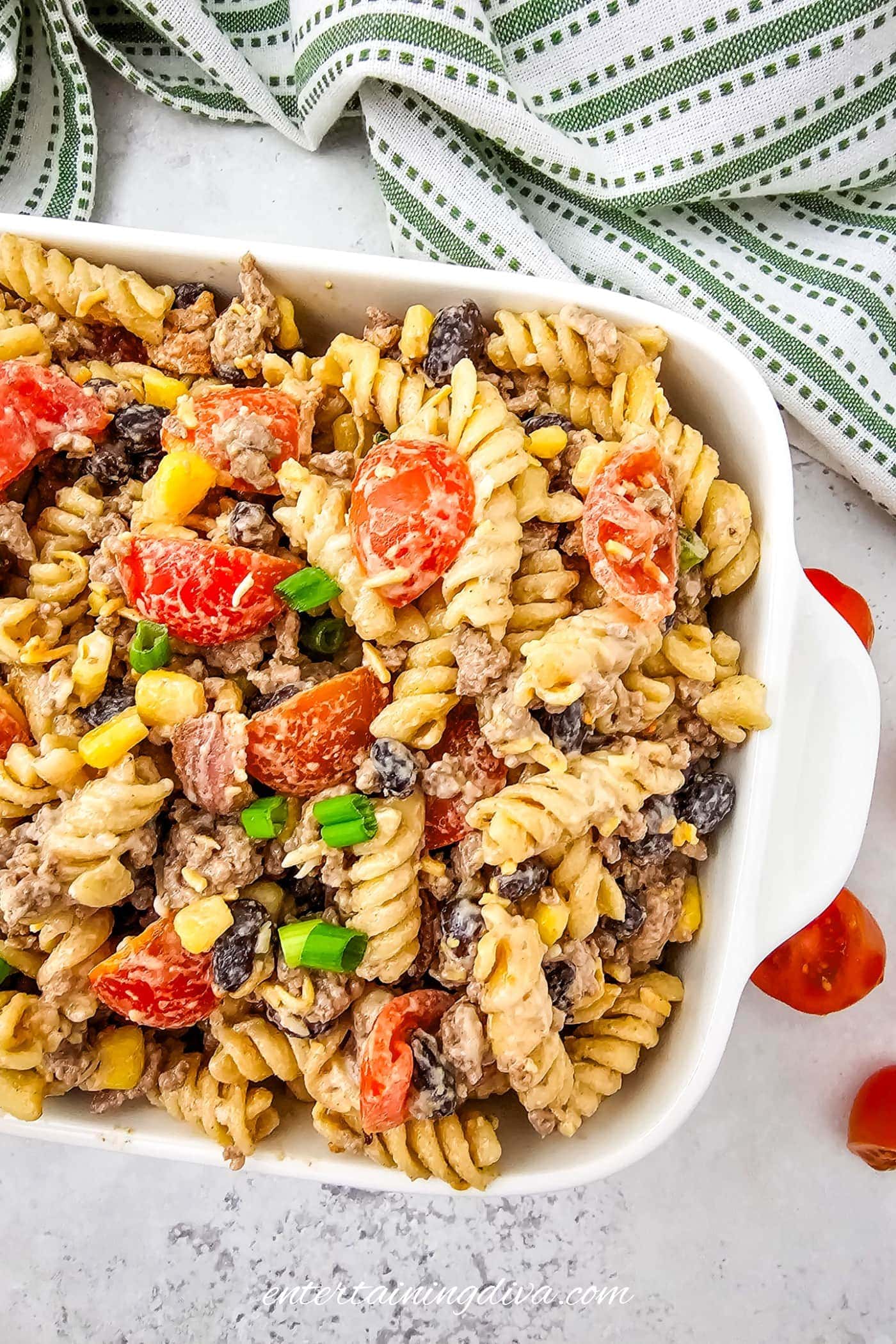 A white casserole dish filled with cowboy pasta salad placed on a light countertop next to a striped kitchen towel.
