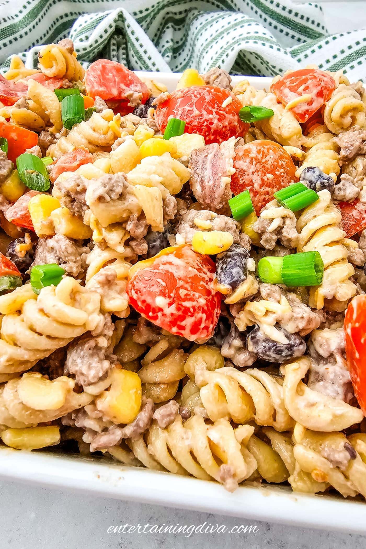 A close-up of a cowboy pasta salad with rotini, ground beef, cherry tomatoes, black beans, corn, and chopped green onions in a white dish.