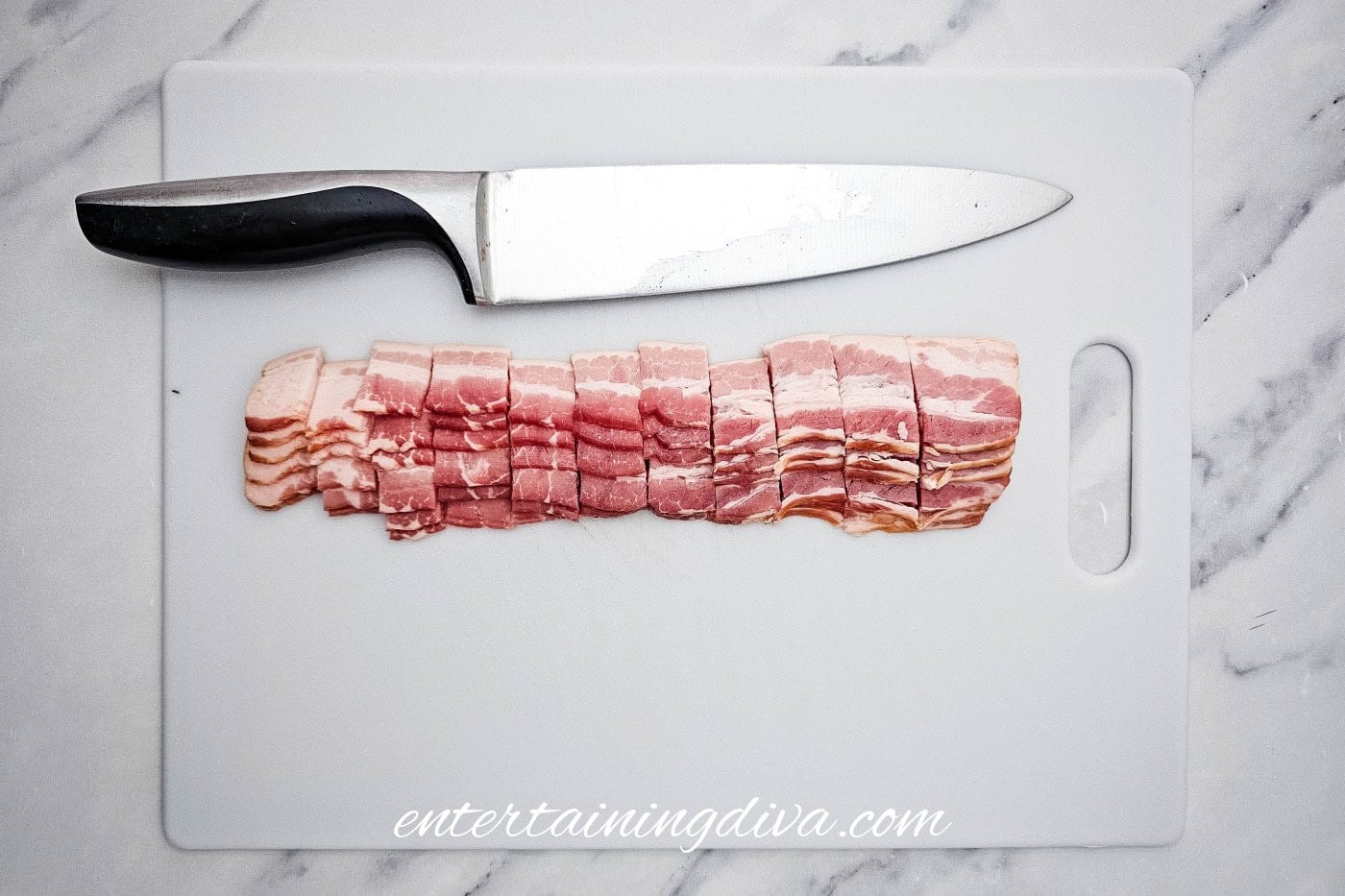 A kitchen knife and several slices of chopped raw bacon on a white cutting board placed on a marble countertop.