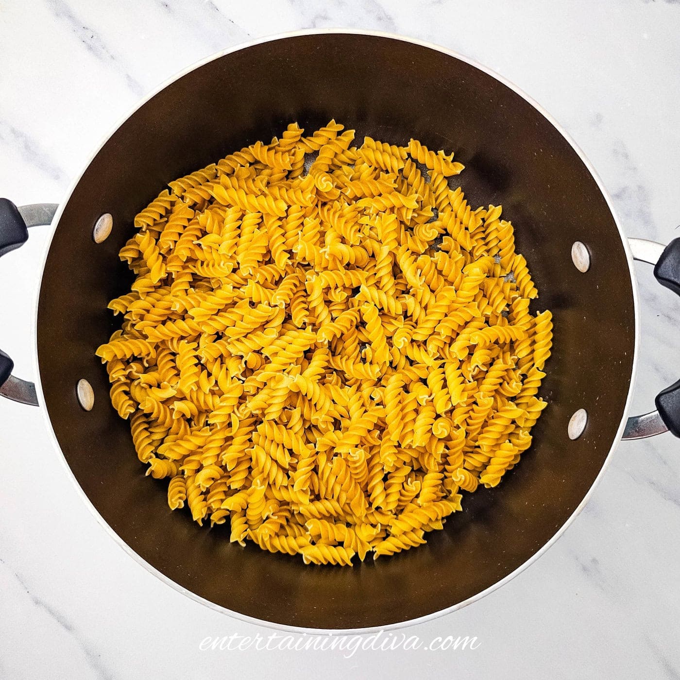 A pot filled with cooked rotini pasta sits on a white marble surface.