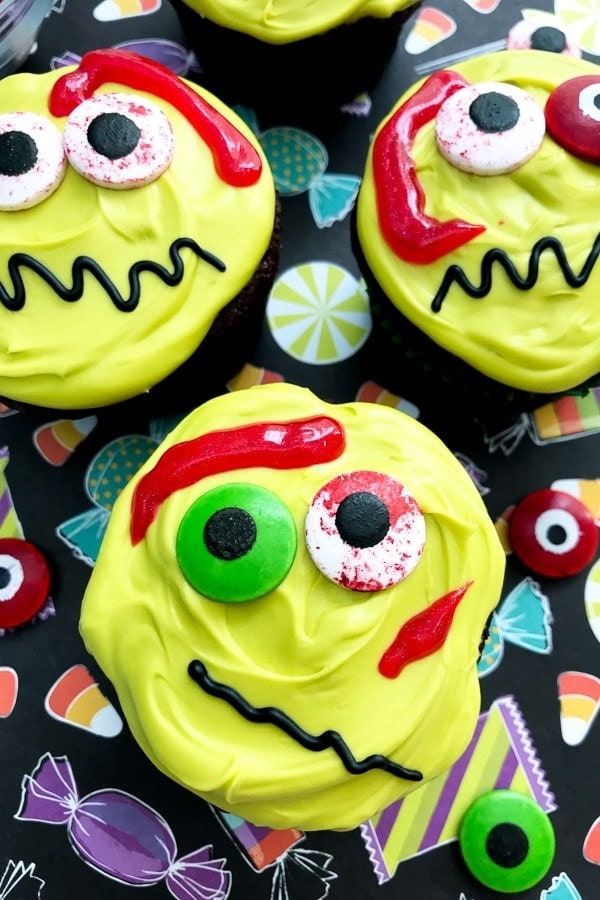 Cupcakes decorated with yellow frosting, candy eyes, red icing, and black icing squiggles to resemble monster faces, placed on a patterned surface with candy illustrations.