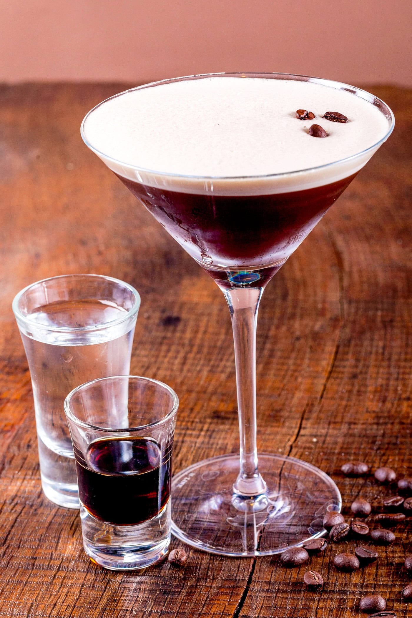 espresso martini garnished with coffee beans beside shot glasses of vodka and coffe liqueur
