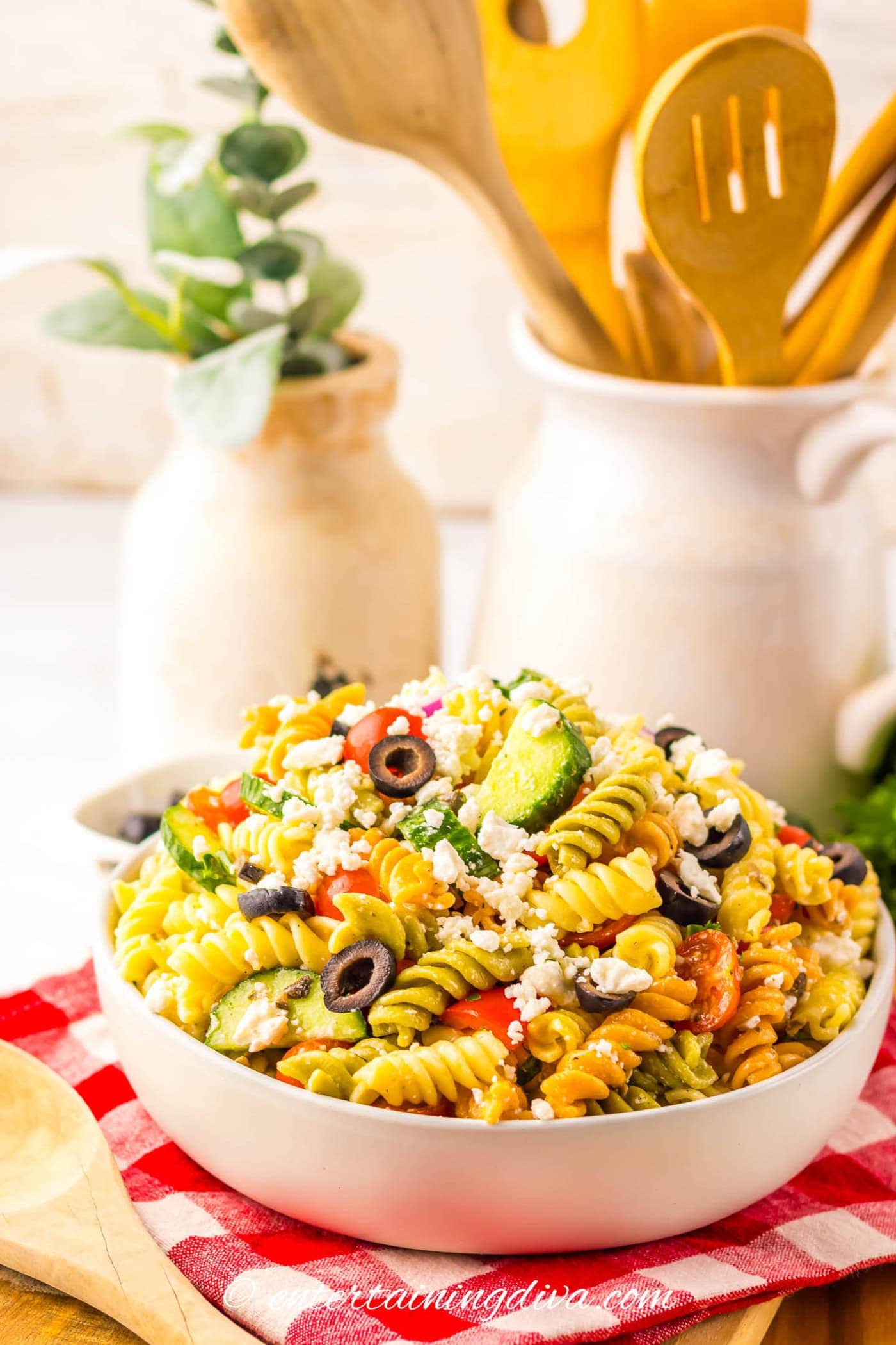 Greek pasta salad in a serving bowl with a container of wooden serving utensils behind it