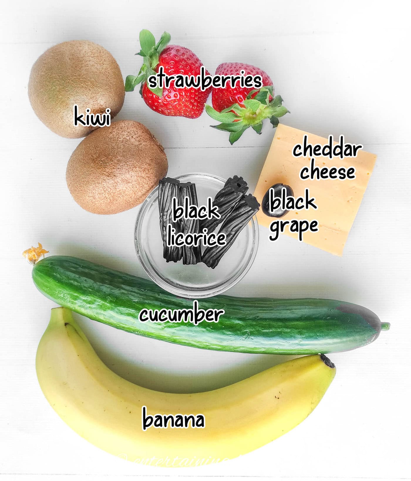 grinch charcuterie board ingredients - kiwi, strawberries, cucumber, banana, black licorice, black grape or olive, cheddar cheese
