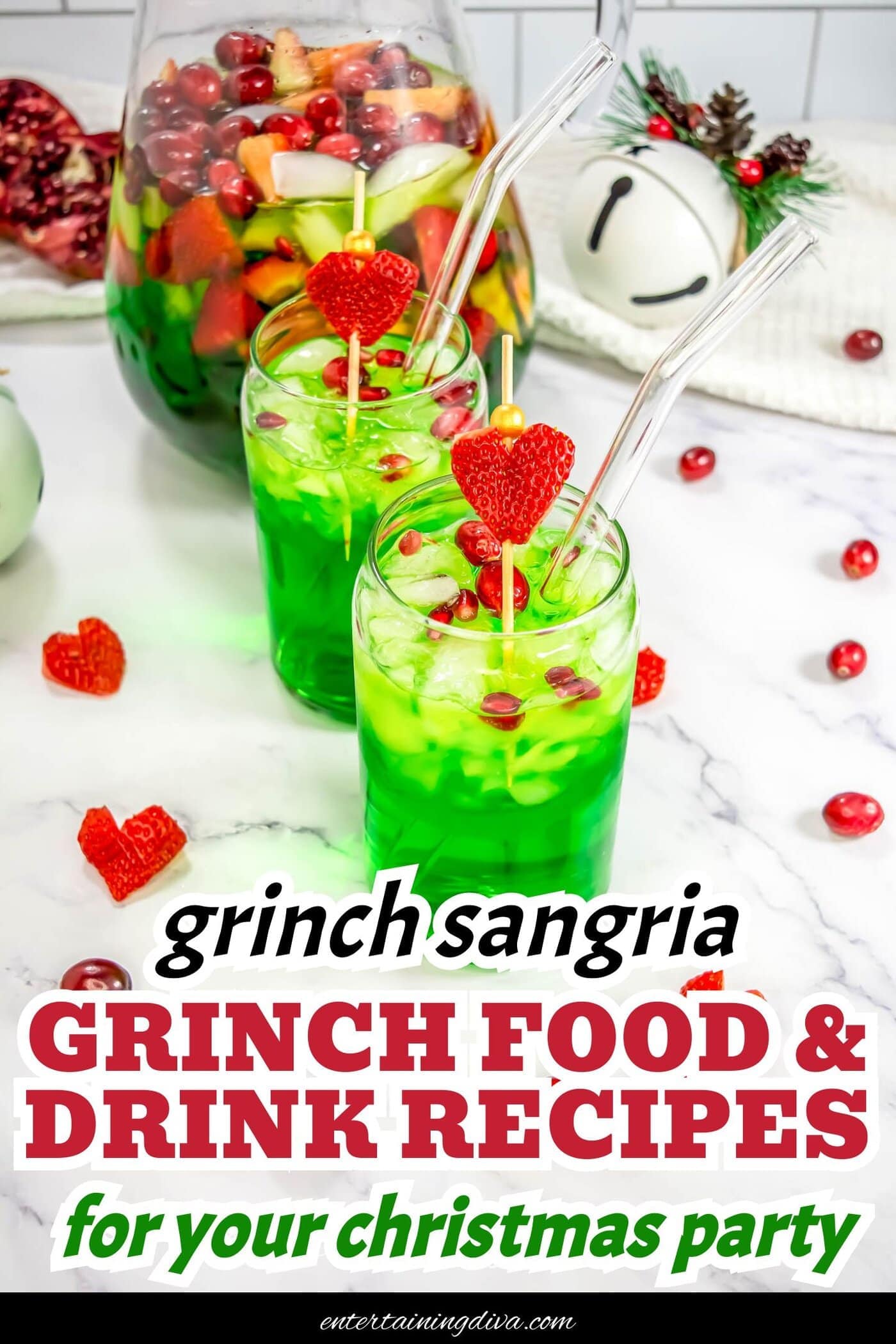 Two glasses of green holiday punch with ice, cranberries, and heart-shaped strawberry garnishes, placed on a marble counter with scattered fruit and a pitcher in the background.