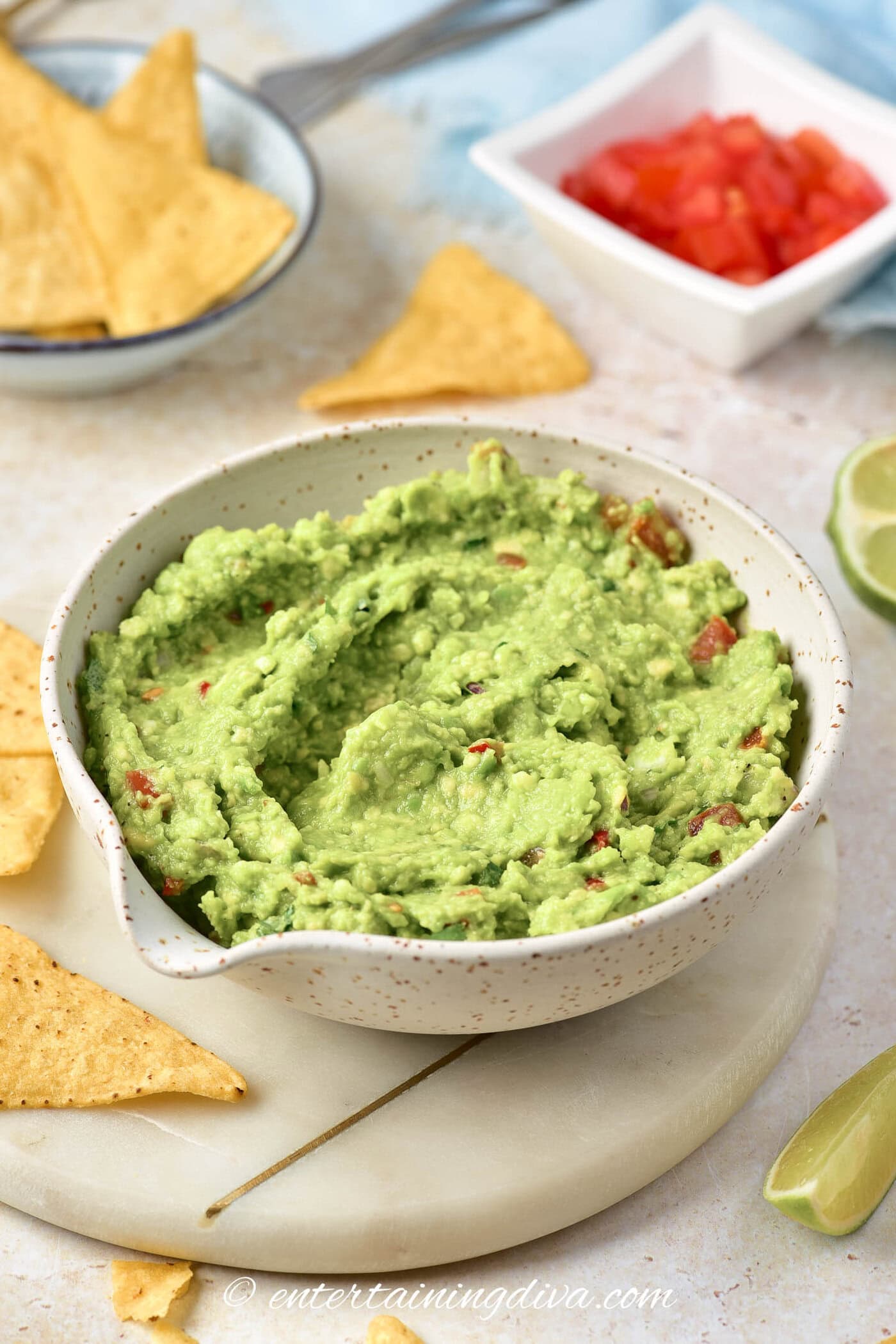 A bowl of guacamole dip with a bowl of tortilla chips, limes and diced tomatoes in the background
