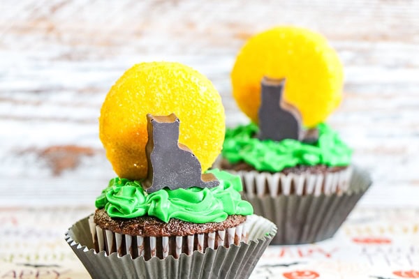 Two chocolate cupcakes with green frosting, topped with a yellow candy disk and a chocolate cat silhouette decoration.