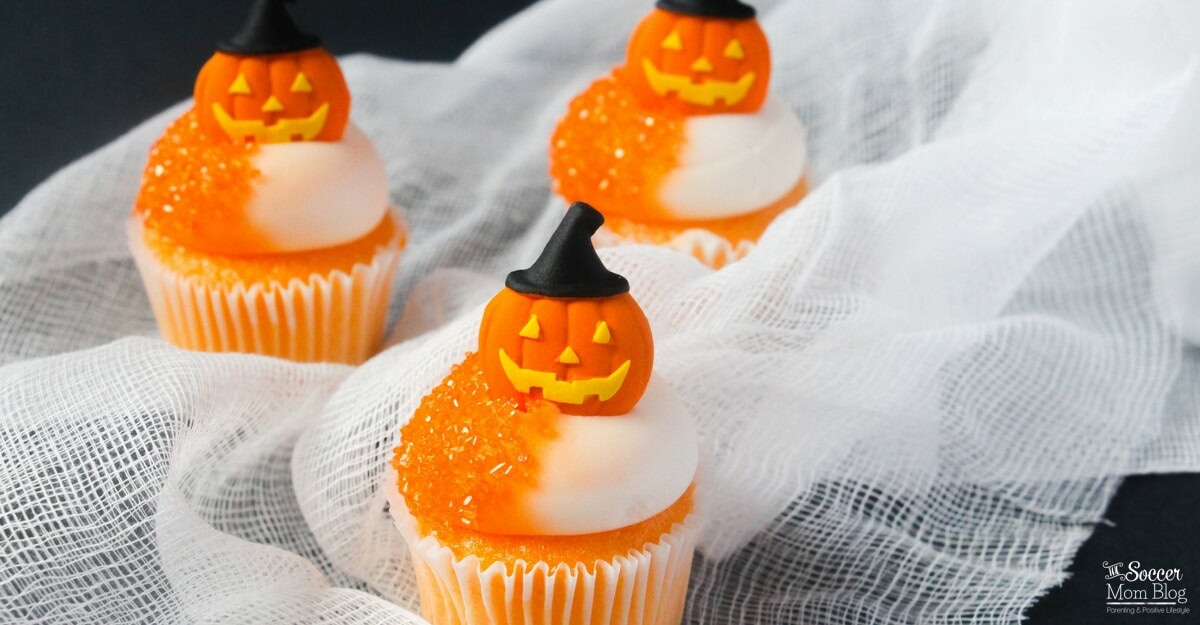 Three cupcakes with white and orange frosting are topped with small pumpkin decorations wearing black witch hats, set on white gauze fabric.