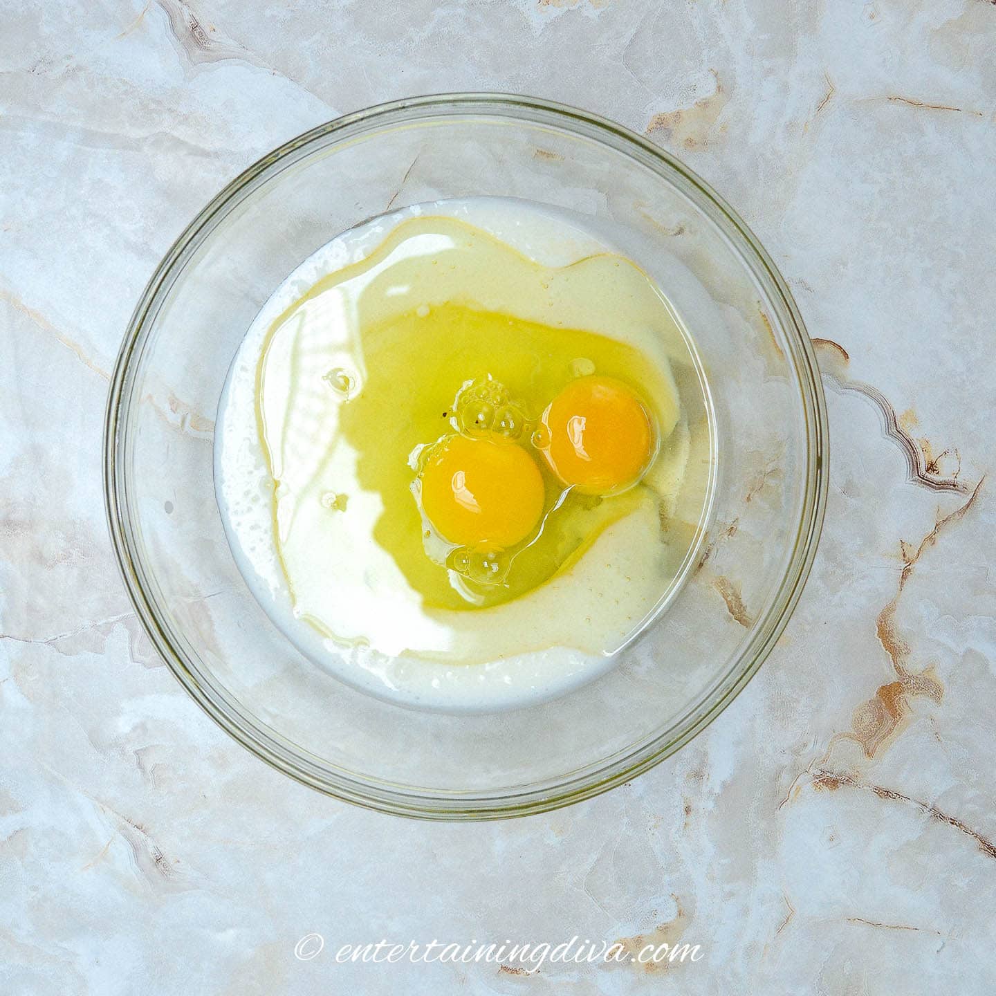Two eggs and other wet ingredients in a glass bowl on a marble surface.