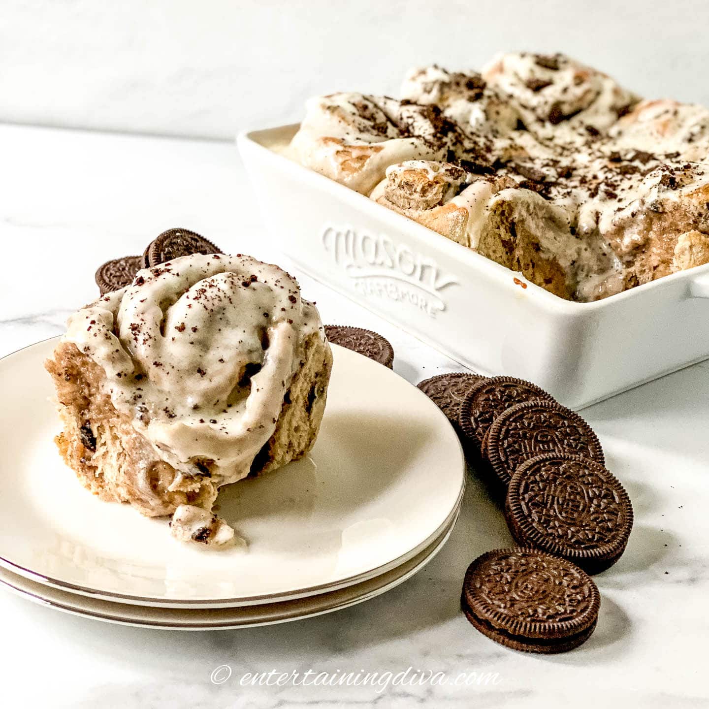 Oreo cinnamon roll with cream cheese filling and vanilla glaze on a plate beside a baking dish full of Oreo cinnamon rolls