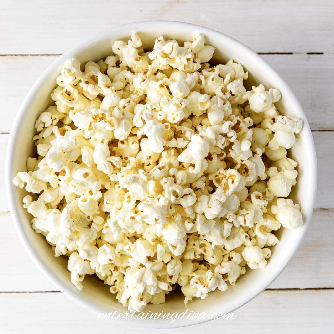 A white bowl filled with plain popped popcorn sits on a white wooden surface.