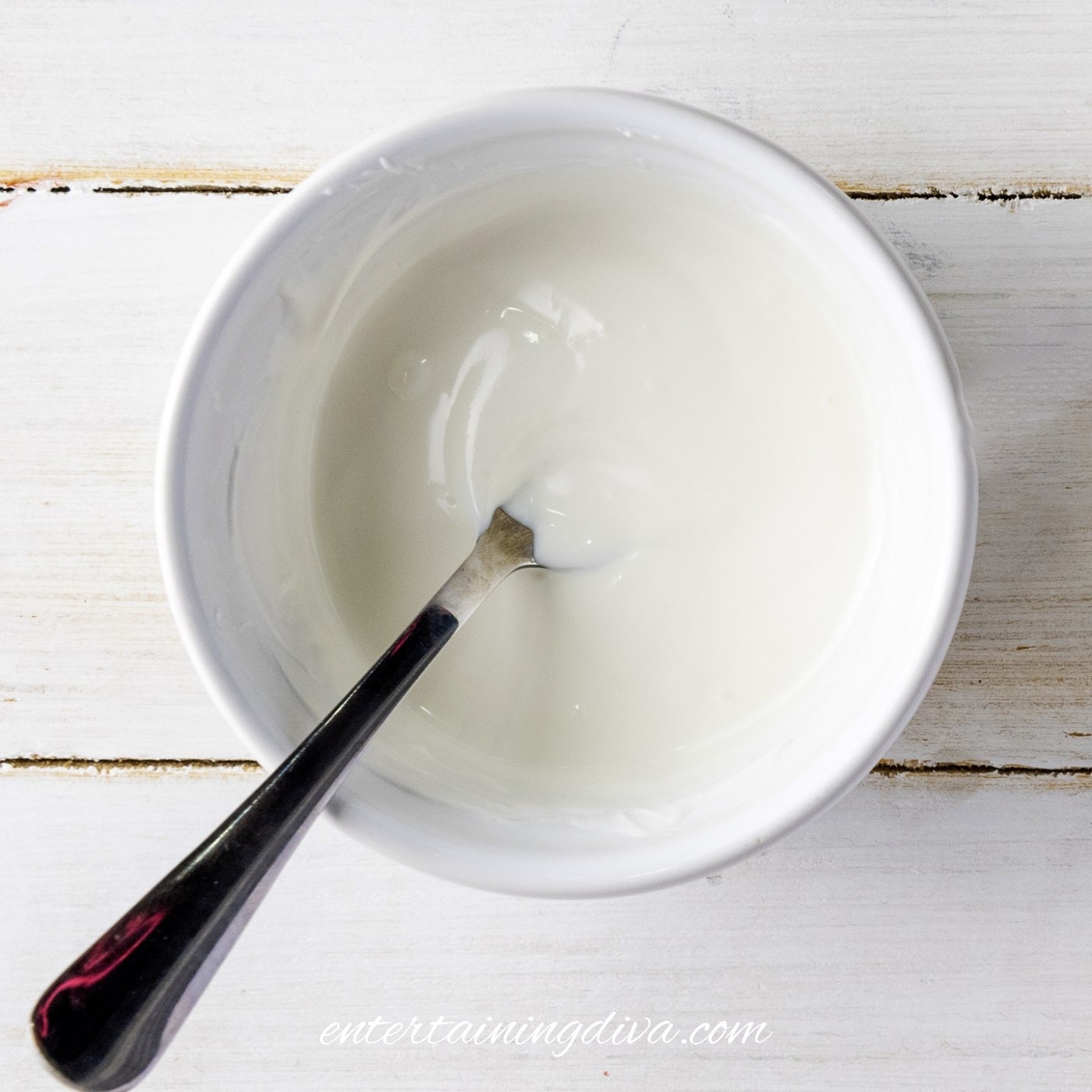 A white bowl with a metal spoon filled with melted white candy melts placed on a white wooden surface.
