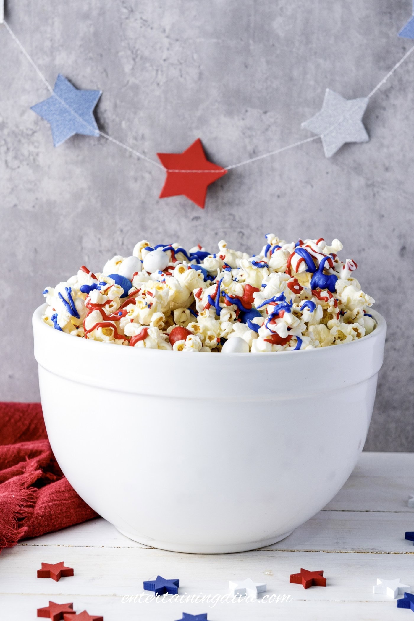A white bowl filled with popcorn topped with red, white, and blue drizzle sits on a white table with star-shaped decorations in the background.