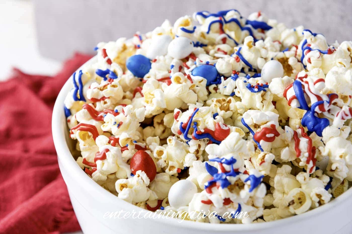 A white bowl filled with popcorn topped with red, white, and blue candy melts and candy pieces, sitting next to a red cloth.
