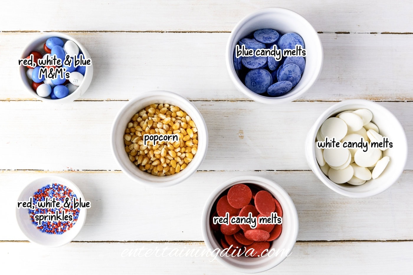 Six white bowls on a white wood surface containing red, white, and blue M&Ms, popcorn kernels, blue candy melts, red, white, and blue sprinkles, red candy melts, and white candy melts.