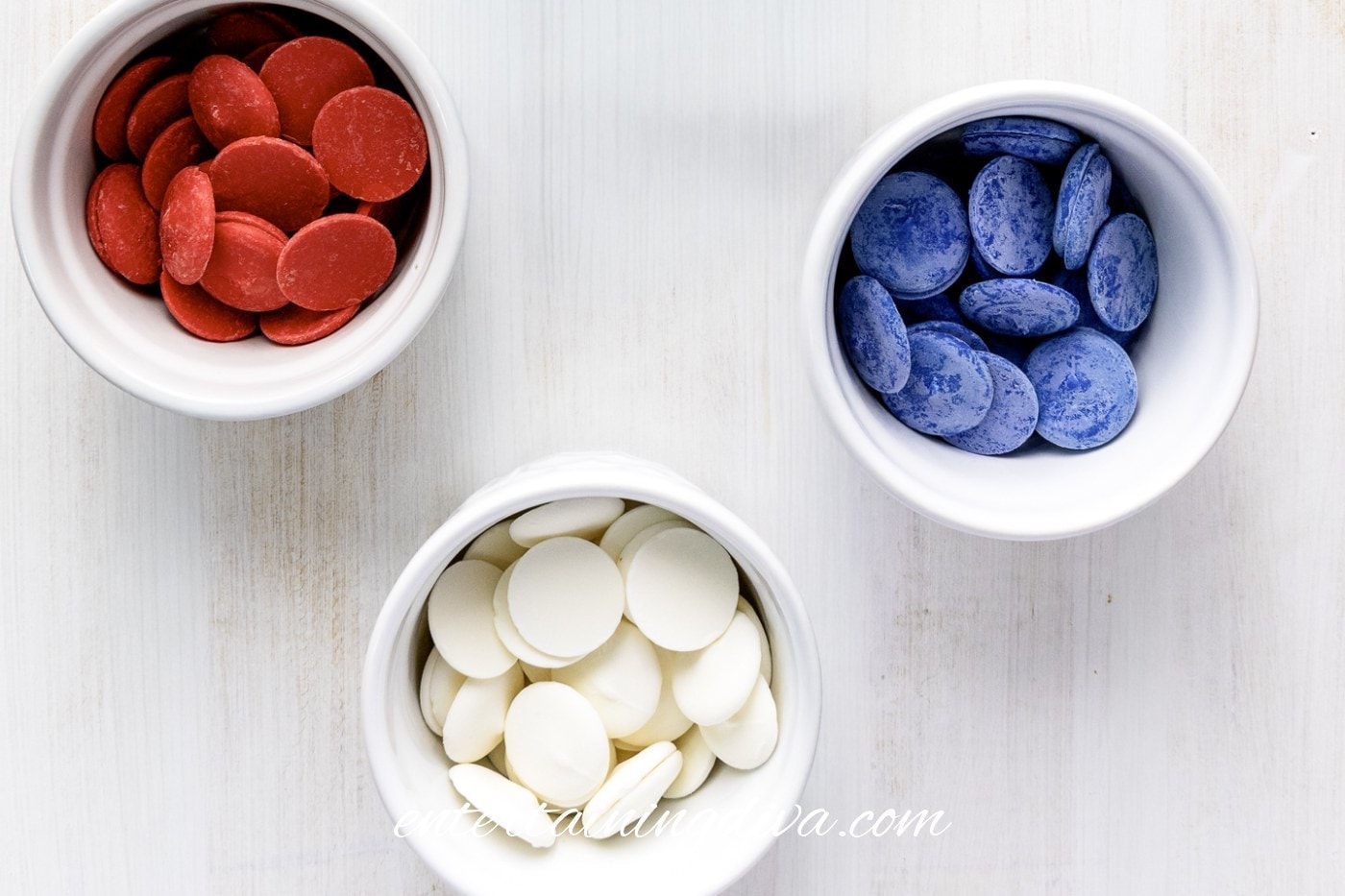 Three white bowls contain red, blue, and white chocolate melting wafers, arranged on a light-colored surface.
