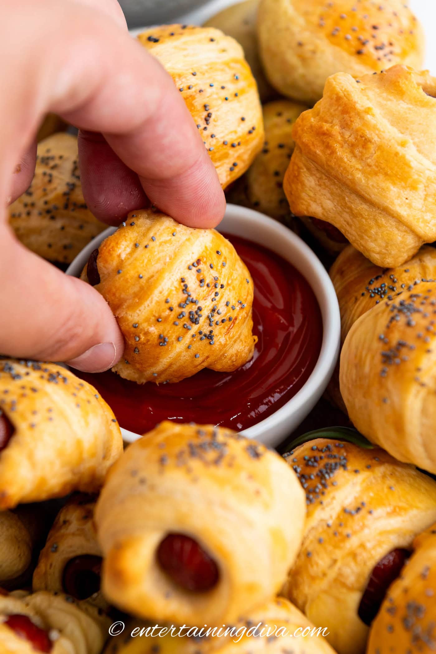one pig in a blanket being dipped in a bowl of ketchup surrounded by more pigs in a blanket