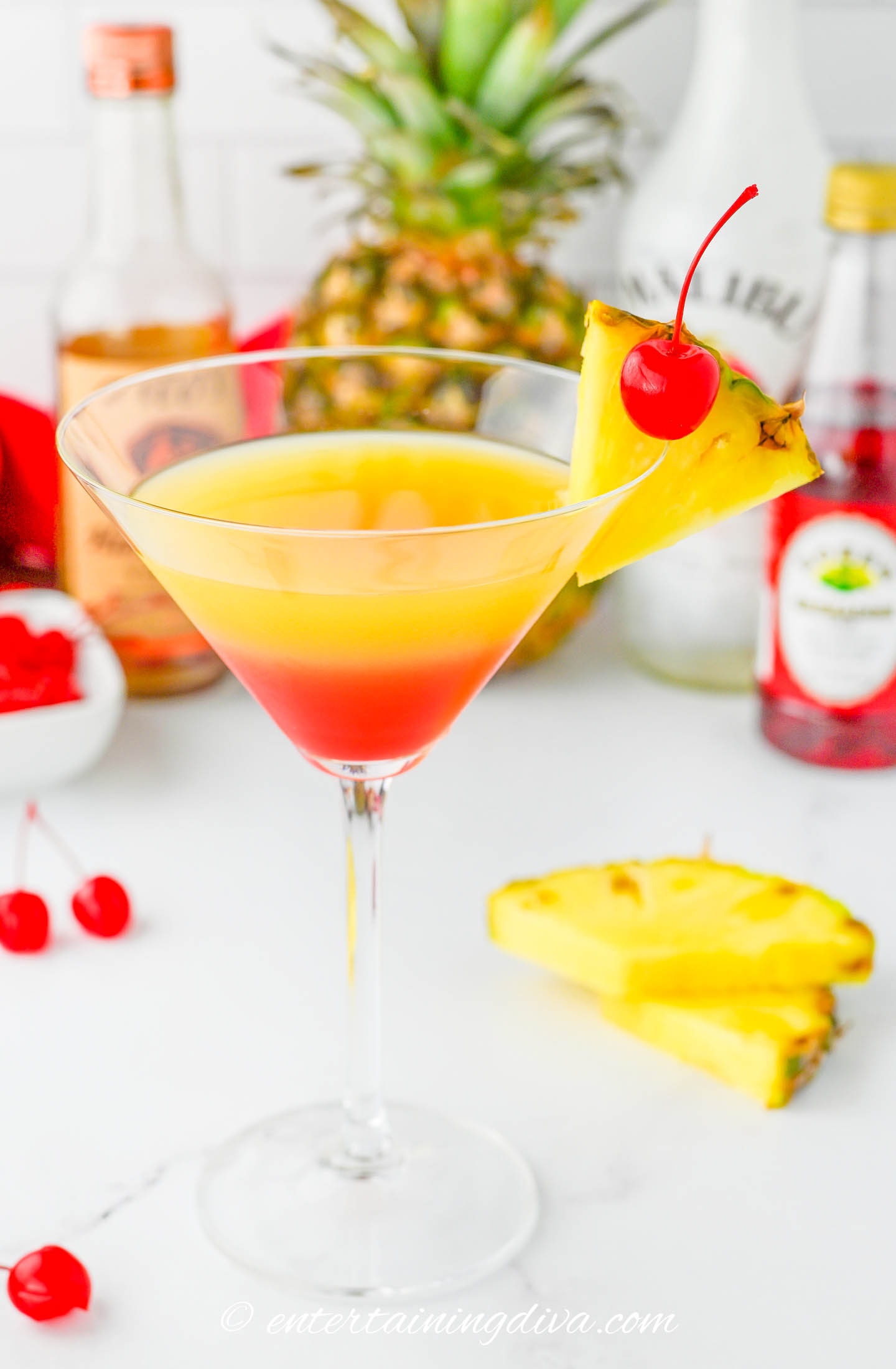 A tequila sunrise cocktail in a martini glass, garnished with a pineapple slice and a cherry, with bottles, pineapple, and cherries in the background.