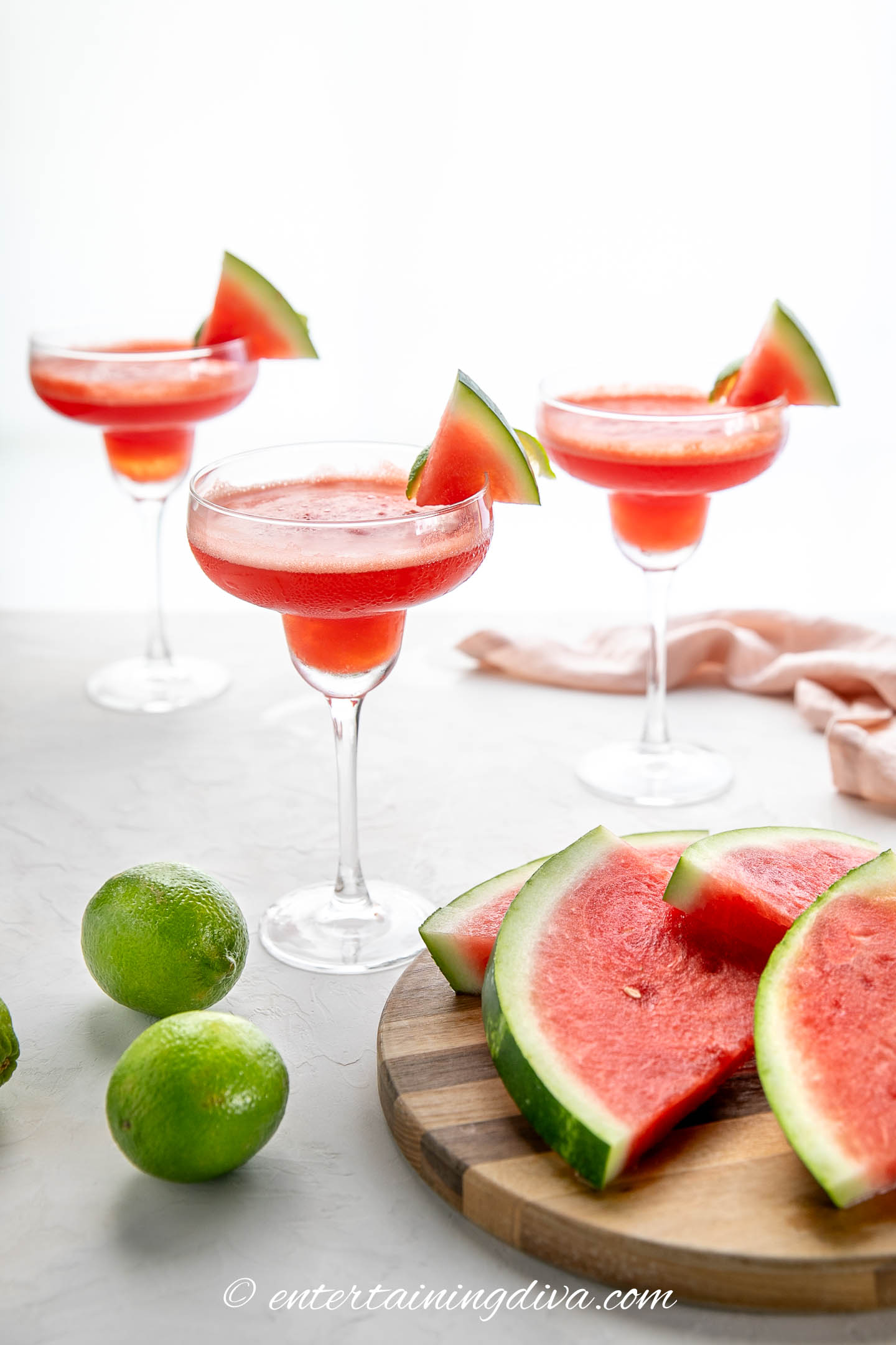 Three watermelon margaritas garnished with watermelon wedges sit on a table with fresh limes and watermelon slices on a wooden board.