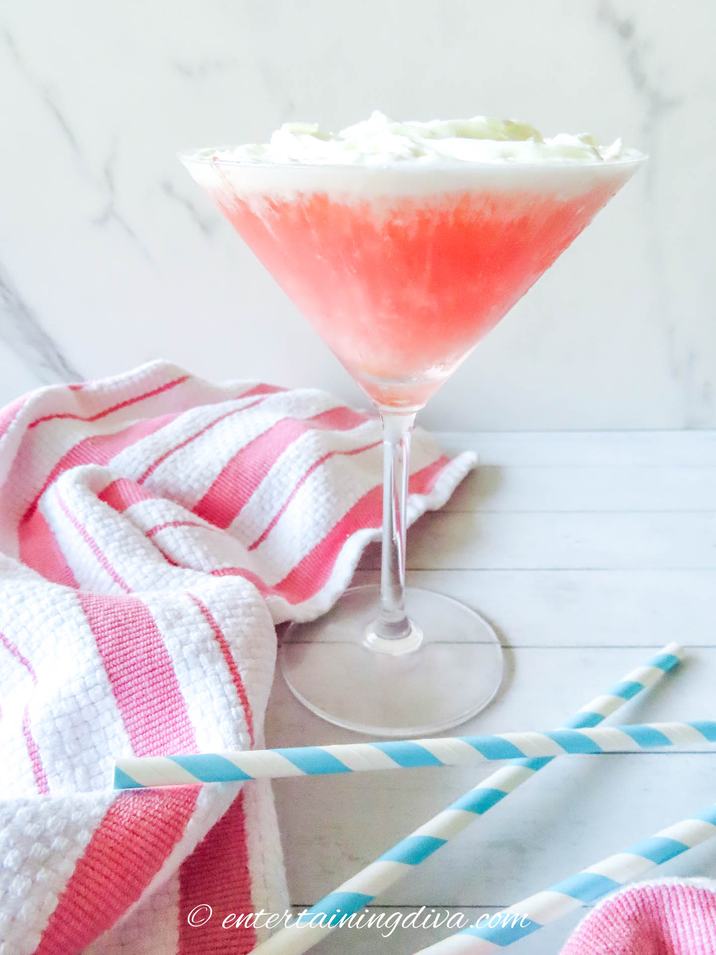 A pink cocktail in a martini glass with a whipped topping, placed on a white surface next to striped straws and a pink-and-white towel.