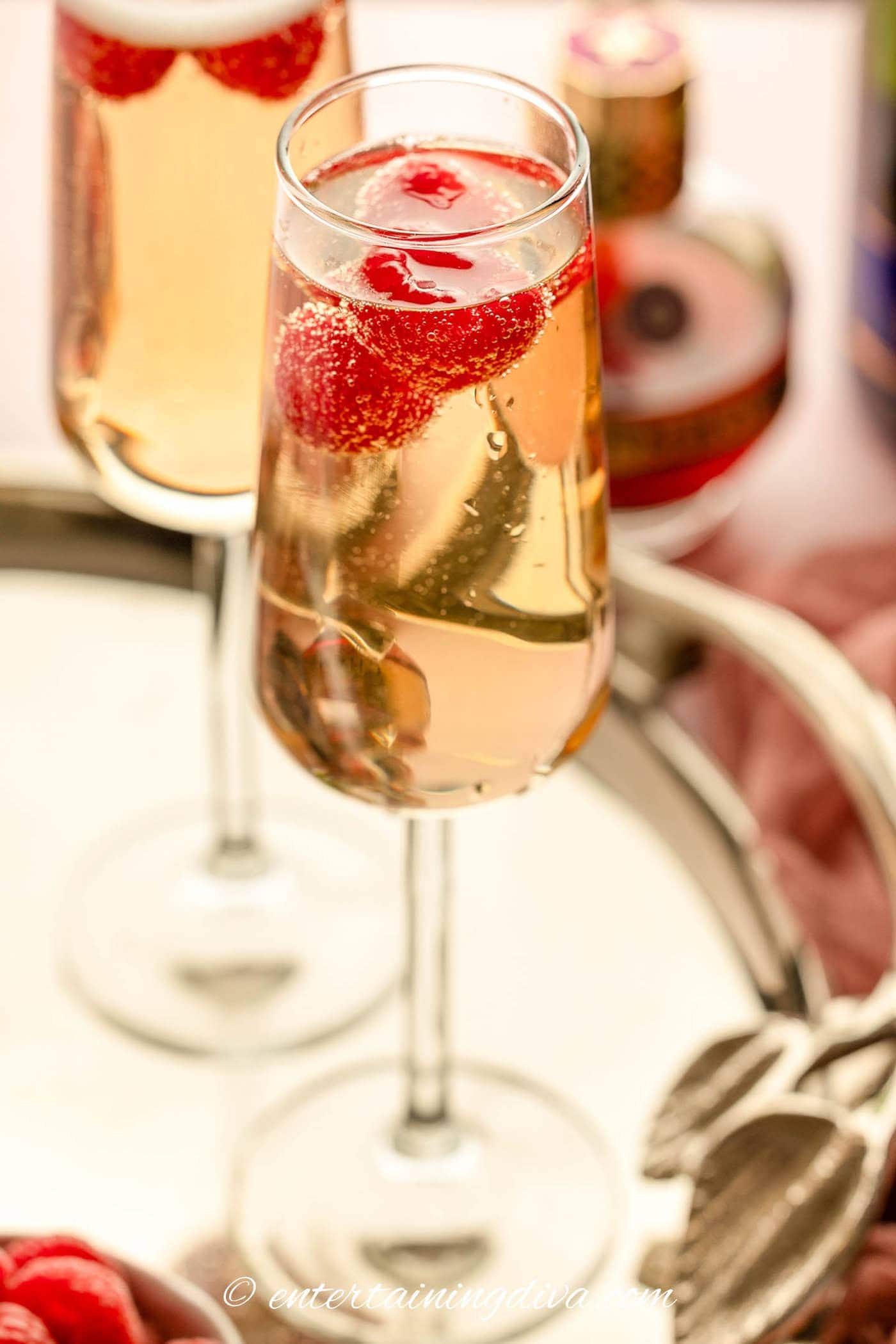 A champagne flute filled with sparkling wine and raspberries sits on a tray, with another similar glass and a bottle in the background.